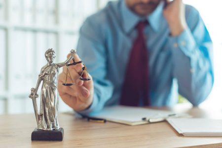 Attorney talking on mobile device and playing with Justice scale in law office, selective focus on statue of Lady Justice